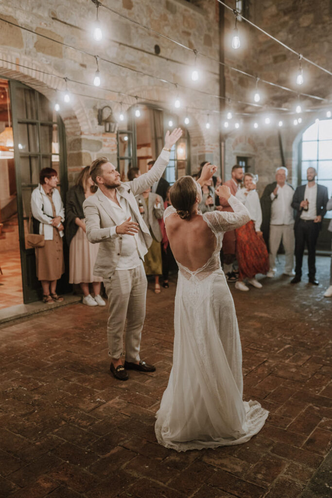 Bride and groom dancing joyfully under string lights at their Tuscany wedding, with guests watching and clapping