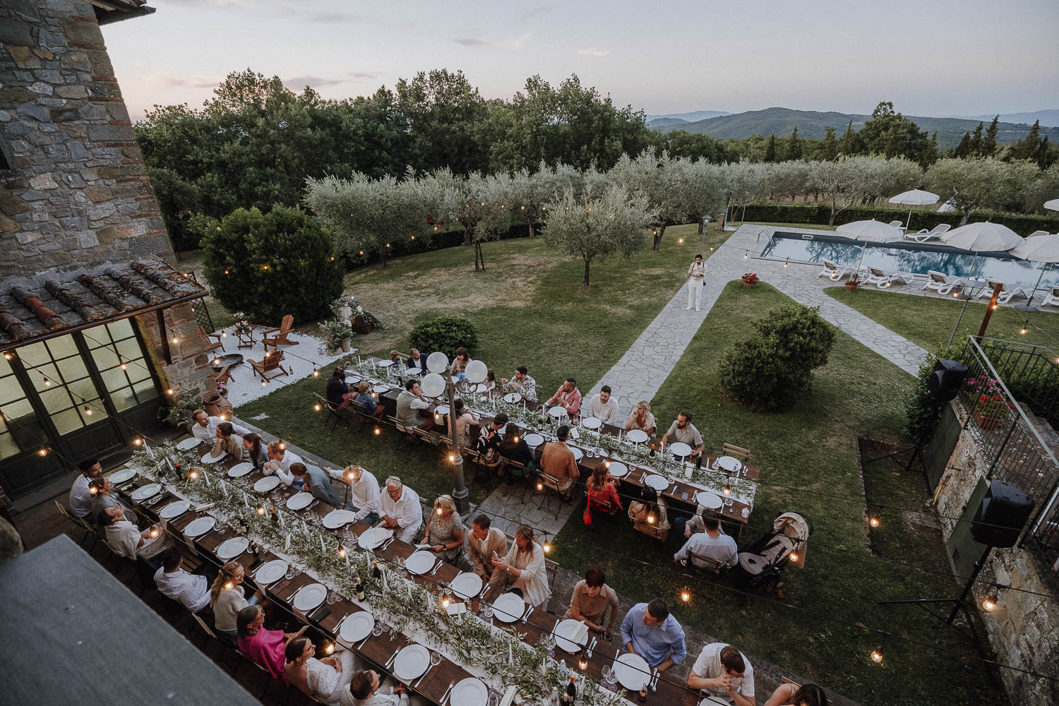 Outdoor wedding dining with guests, elegant tables, string lights, stone building, garden, and pool at sunset.